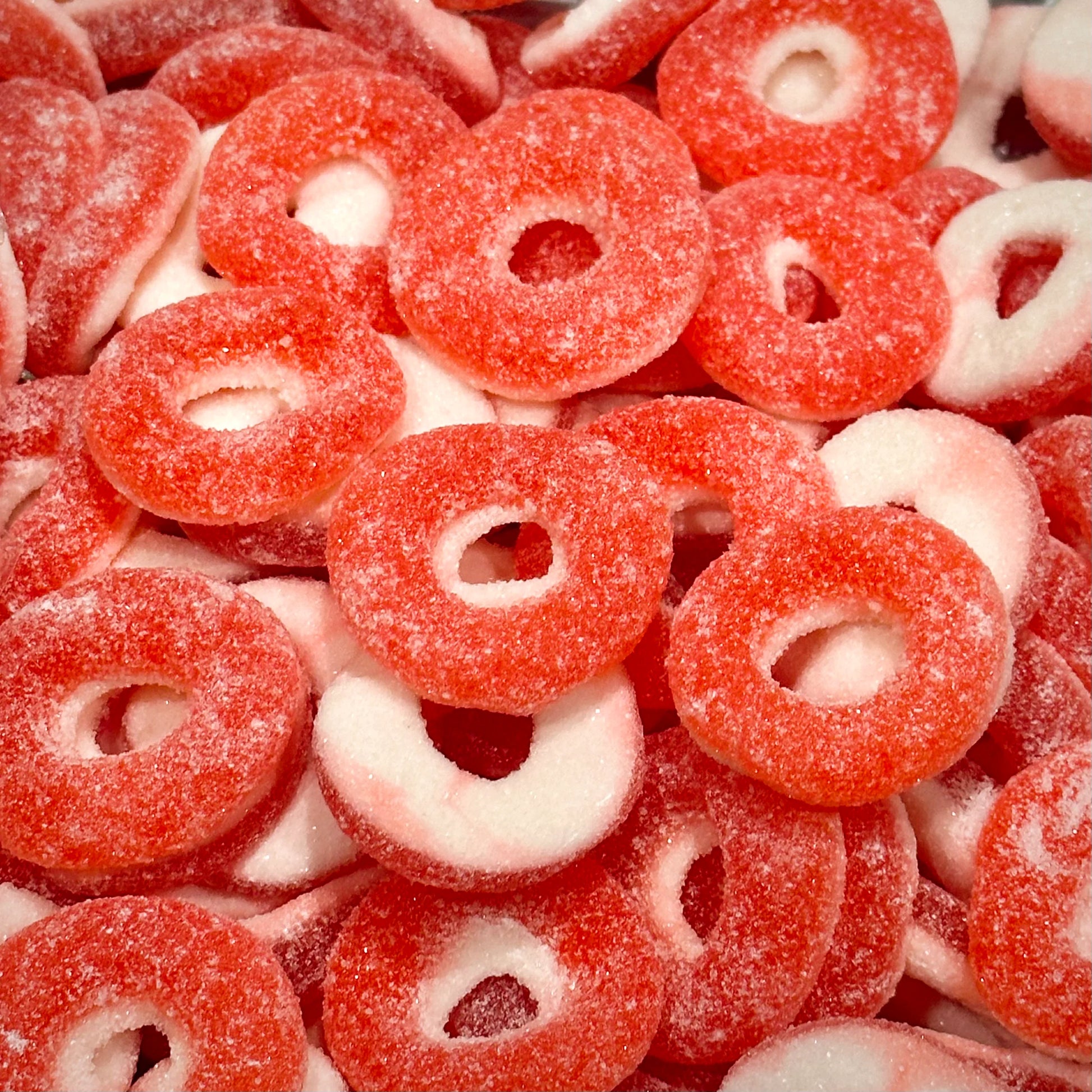 Red and white strawberry ring gummy candies covered in sugar crystals, showing circular ring shapes with hollow centers scattered across the frame